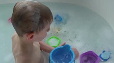 Küçük çocuk köpük dolu bir küvette banyo yaparken oyun oynuyor. Banyo oyuncaklarıyla çevrili çocuk, banyo zamanının neşeli atmosferini kucaklayarak odaklanmış ve rahatlamış görünüyor.. 