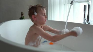 Çocuk küvette saç yıkıyor, sabunlu köpüklerle kaplı, musluktan akan su. Sahne banyo zamanındaki neşeli ve eğlenceli anı yakalıyor, temizlik ve eğlenceyi vurguluyor.. 