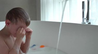 Küçük bir çocuk, beyaz oval küvette banyo yapmaktan zevk alıyor, kafasını musluktan akan su deresinin altına eğiyor ve sonrasında gözlerini siliyor.