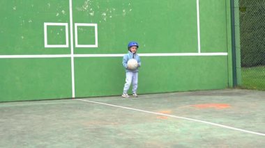 Çitli tenis kortunda sessiz bir sahne. 4 yaşında bir çocuk futbol oynuyor, beyaz bir topa vuruyor ve top çimlere uçuyor.