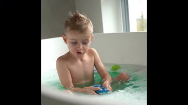 Çocuk geniş bir küvette turkuaz suyla banyo yapıyor, oyuncaklarla oynuyor, oda, yerden tavana geniş pencereler sayesinde güneş ışığıyla dolu. Yüksek kalite 4K görüntü. Yüksek