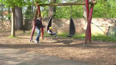 Ebeveynler ve çocuklar oyun parkında eğleniyorlar. Çocuk, yetişkin yanında dururken salıncakta mutlu bir şekilde oturur. Arkaplanı ağaçlar ve ahşap yapılar oluşturuyor..