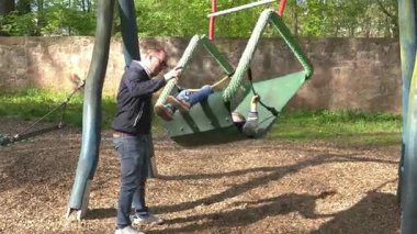 Çocuk, çocuk parkındaki koltukta sevinçle sallanırken bir yetişkin de çocuğun güvenliğini sağlamak için etrafı kolaçan ediyor. Yeşil ağaçlar ve taş duvar zeminiyle güneşli bir gün..
