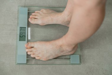 Bireyler, kilolarını gösteren görünür bir ekran ile dijital ölçekte yalın ayak dururlar. Ayar, bir anlık kendini ölçmenin vurgulandığı fayanslı bir zemin gösteriyor..