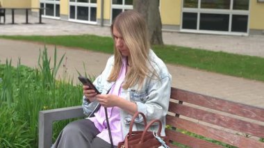 Parktaki bankta oturan genç bir kadın dikkatle akıllı telefonuna bakıyor. Günlük kıyafetleri arasında bir kot ceket ve çevresi yeşilliklerle çevrili. Açık havada sakin bir ortama dikkat çekiyor..