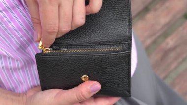 Altın paraları ortaya çıkarmak için siyah bir cüzdan açarken görülen eller cüzdan detaylarına ve el hareketlerine odaklanıyor. Açılan cüzdanın dokusu ve tasarımı vurgulanır.