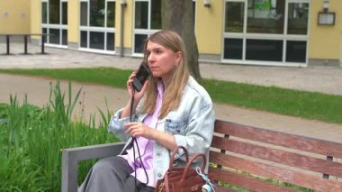 Parkta bankta oturan bir kadın telefonuyla konuşuyor. Konuşmasına odaklanmış ve meşgul görünüyor. Kot ceket giyiyor ve etrafı barışçıl park yerleriyle çevrili..