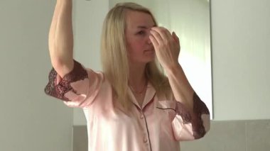 Banyoda pembe pijamalı bir kadın saçına saç spreyi sıkmış. Spreyden korunmak için yüzünü kapatıyor. Kişisel bakım ve saç şekillendirme rutini.