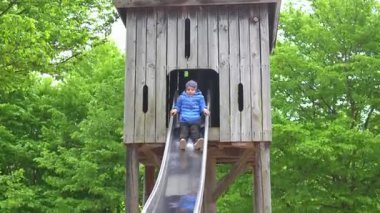 Mavi ceketli çocuk tahta bir oyun parkından kayıyor. Etrafı yemyeşil ağaçlarla çevrili çocuk, neşeli bir ifade sergiliyor, dışarıda eğlenme ve oyun oynama deneyimi yaşıyor..