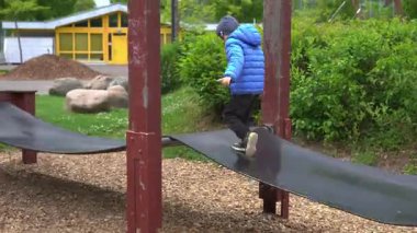 Mavi ceket giyen bir çocuk parkta oyun parkı ekipmanlarında denge sağlar. Hareket ve açık hava eğlencesi odaklı aksiyon, oyun ve macerayı yakalar. Arka planda yeşillik ve sarı bina