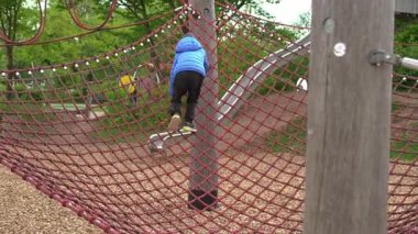 Mavi ceket giyen çocuk oyun parkında kırmızı ağa tırmanıyor. Açık hava oyun alanında yeşil ağaçlar ve bir slayt, çocuklar için aktif oyun ve keşif teşvik edici nitelikte..