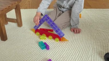 Çocuk halıya oturmuş renkli yapı taşlarıyla oynuyor, basit bir yapı oluşturuyor. Elleri blokları düzenliyor, yaratıcılığı ve motor becerileri teşvik ediyor..