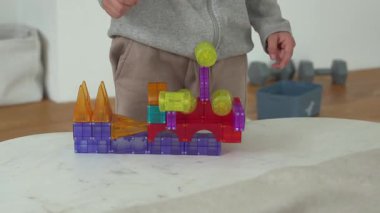Çocuk, kapalı bir masada renkli manyetik bloklarla yaratıcı bir şekilde oynuyor. Eller aktif olarak canlı geometrik şekilleri düzenlemekle, hayal gücünü ve problem çözme becerilerini teşvik etmekle meşgul..