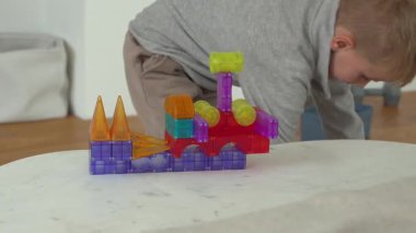 Beyaz bir masanın üzerinde renkli bir blok kulesini birleştiren çocuk. Yapıda çeşitli şekiller ve canlı renkler yer alıyor. Eller, kapalı alanda oynanan oyunda bağlılık ve yaratıcılığı öneriyor..