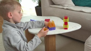 Masanın üzerinde renkli manyetik bloklarla yaratıcı bir oyun oynayan küçük bir çocuk, odak ve merakla şekiller oluşturuyor. Arka planda rahat mobilyalar olan kapalı bir ortam..