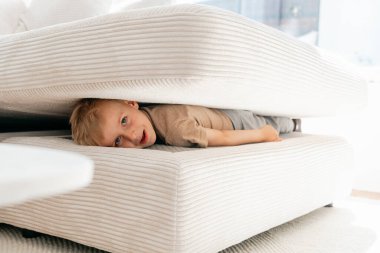 Çocuk beyaz bir kanepenin yastıklarının arasına saklanmış, tuhaf ve yaramaz bir ifade sergiliyor. Samimi ortam eğlenceli ve yaratıcı bir oyun anı olduğunu gösteriyor..