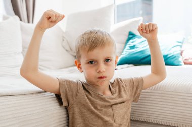 Kararlı bir ifadeye sahip çocuk, kolları bükülmüş, yastıklı, modern bir kanepeye oturmuş güven içinde poz veriyor. Ev ortamında rahat ve rahat bir atmosfer vurgulanıyor