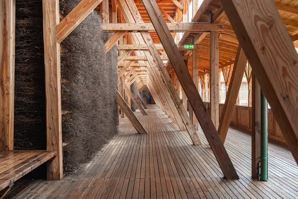 Büyük kirişler ve doğal malzemelerle ahşap mimari iç mekan eşsiz bir geçit oluşturuyor. Bir kişi arka planda duruyor ve geometrik tasarıma ölçek ekliyor. Tuz Mezuniyet Kulesi