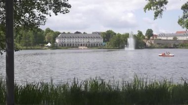 Arka planda yemyeşil bir göl ve tarihi binaların olduğu huzurlu bir manzara. İnsanlar sakin sularda dinlendirici bir pedal sürüşünün keyfini çıkarıyorlar. Huzurlu ve resimsel bir ortam yaratıyorlar.