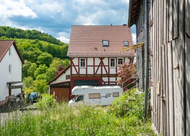 Güneşli bir günde yemyeşil bir araziye kurulmuş, yakınlarda bir karavan park etmiş, yarı keresteli sevimli bir ev. Geleneksel mimari, huzurlu bir kırsal alanda modern seyahatle karşılaşıyor..