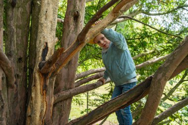 Çocuk, açık renk ceket ve kot pantolon giyerek büyük bir ağaca tırmanır. Macera ve merak hissi uyandırır. Yoğun yeşillik çevresi, doğal ortamı vurgular..