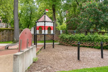 Kale temalı bir yapıya sahip renkli bir oyun alanı, yemyeşil bir park ortamında kaydırak ve ip köprüsü var. Çocuklar için açık hava aktiviteleri ve yaratıcı oyunlar için ideal..
