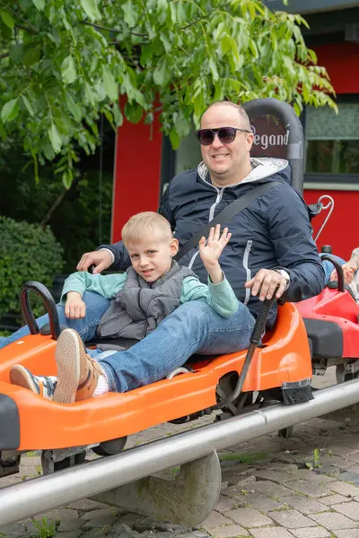 Homberg, Almanya, 06.09.25. Baba ve oğul, turuncu bir hız treninde oturmuş, keyifli bir anı paylaşıyorlar. Çocuk kameraya gülümseyerek el sallıyor, eğlence parkının heyecanını yakalıyor.