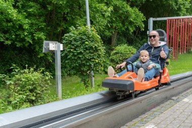 Baba ve oğul, bereketli yeşilliklerle çevrili bir lunapark treninde keyifli bir anı paylaşıyorlar. Çocuk heyecanla gülümserken baba başparmak işareti yapıyor..