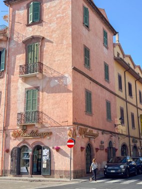 Bosa Italia 24.06.25 şehir manzarasında karşıdan karşıya geçen bir kadın süslü balkonları ve panjurları olan eski bir İtalyan binasının önünden geçiyor. Açık gökyüzü binaları güzelleştiriyor..