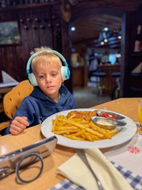Kulaklık takan genç bir çocuk, bir tabak patates kızartması ve pirzola ile ahşap bir masada rahat bir restoran ortamında oturuyor. Odaklanmış görünüyor, muhtemelen bir cihazdan bir şey izliyor.