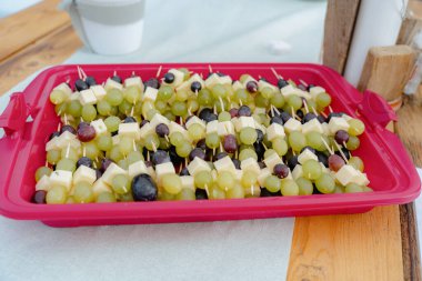 Lezzetli peynir ve üzüm şişleri canlı kırmızı bir tepside düzenlenir. Parti ve atıştırmalıklar için idealdir. Yeşil ve siyah üzümler peynir küpleriyle eşleştirildiğinde görsel olarak çekici bir tabak oluştururlar..