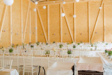 Beyaz çarşaf ve sandalyelerle süslenmiş, çiçeklerle süslenmiş masaların olduğu bir ziyafet salonu. Kırsal ahşap duvarlar ve asılı süslemeler zarif atmosfere ekleniyor..