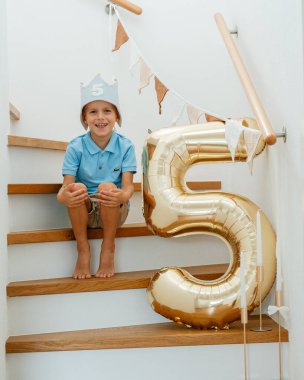 Beş yaşında neşeli bir çocuk elinde büyük altın bir balonla merdivenlerde oturmuş, evinde doğum gününü şenlikli bir dekorla kutluyor.