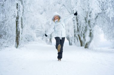 Kış kıyafetleri giyen bir kadın, ormandaki karla kaplı bir patikada neşeyle dans ediyor. Parlak bir gülümseme ve enerjik bir poz serin, soğuk ağaçların arasında mutluluğu iletir.