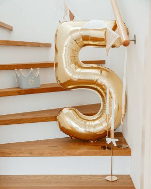 Tahta bir merdivenin üzerinde duran 5 numaralı büyük altın balon bayramlık bir kutlamayı ya da doğum gününü işaret ediyor. Yakınlarda bir taç süslemesi ve mumlar kutlama temasına eklenir..