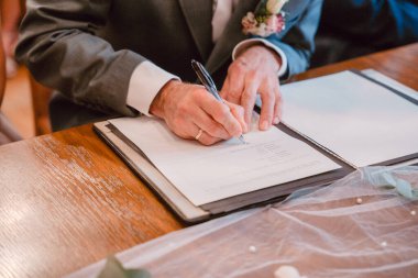 Resmi kıyafetli bir insanın düğün belgesini imzalaması. Görüntü, ahşap bir yüzeye karşı kurulmuş bağlılığı ve hukuki birliği simgeleyen anlamlı bir anı yakalıyor.