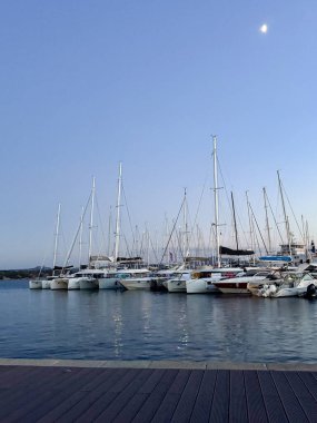 Yelkenliler, ayın aydınlattığı sakin bir akşam gökyüzüne karşı bir marinada dizilir. Sakin su tekneleri yansıtıyor, alacakaranlıkta resim gibi bir manzara yaratıyor..