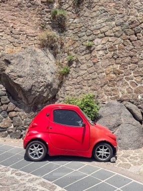 Kentsel bir ortamda, dokulu taş bir duvara park edilmiş kırmızı kompakt araba. Canlı kırmızı renk, doğal taş zemin ile çelişiyor ve eşsiz bir görsel sahne yaratıyor..