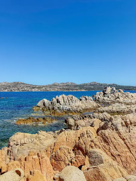 Engebeli kıyı kayaları açık mavi gökyüzünün altında kıyı boyunca uzanır. Deniz turkuvaz suları kayalık kıyı şeridiyle buluşur ve uzak tepelerle çarpıcı bir doğal manzara yaratır.