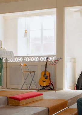 Katlanan sandalye ve nota sehpasının yanında duran akustik gitarlı güneşli bir oda. Ayarlar, müzik alıştırması veya performans için ideal barışçıl bir atmosfer olduğunu gösteriyor.