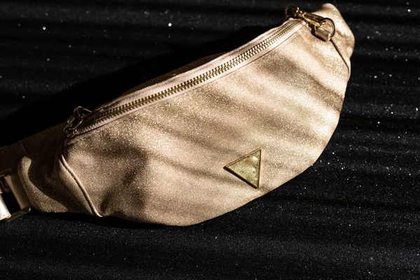 Glamorous womens waist bag in gold color on a black shiny background. Fashionable handbag with gold chain.