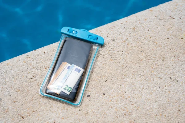 Waterproof case with money and a smartphone on the background of the pool on a sunny summer day.