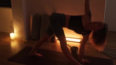 Bir kadın loş ve sıcak ışıkta evde yoga yapıyor. Derin nefes al ve konsantre ol..