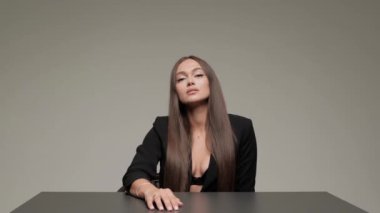 İş ceketli bir kadın masaya oturur ve gri arka planda stüdyoda ciddi bir şekilde kameraya bakar..
