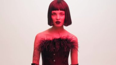 Fashion portrait of a girl in a black bob wig and black lipstick in red light on a white background. Black leather gloves. The girl shows the fuck