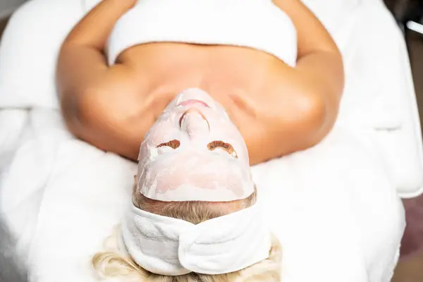 Blonde woman in a salon with a fabric moisturizing mask on her face. Caring for healthy skin.