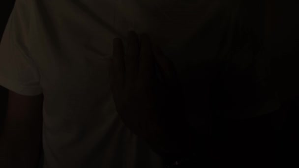 一个穿着白色T恤的男人在一间黑暗的房间里把他的手放在胸前 随着你的心跳节奏闪烁着光芒 中风和心脏病的症状 — 图库视频影像