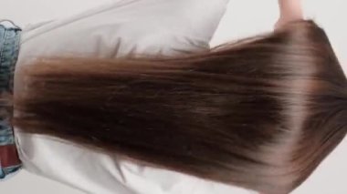 Dikey video. Stüdyoda gri bir arka plana karşı duran esmer bir kadın arkasında duruyor ve saçını büyütüyor. Güzel saçlarla ilgilenmek.