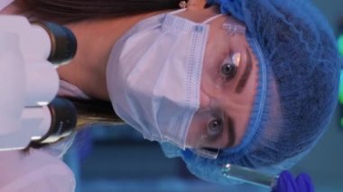 Kadın bir laboratuvar asistanı, maskeli, gözlüklü ve tıbbi şapkalı bir virüs taşıyan bir deney tüpüne bakıyor. Dikey video.