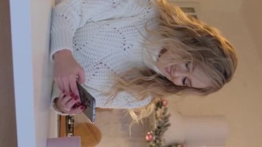 Beyaz kazaklı güzel sarışın bir kadın akıllı telefona bakıyor ve gülümsüyor. Dikey video.